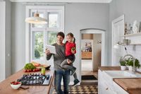 Haustechnik per Tablett steuern, das geht. Beispiel-Foto zeigt Vater mit Kind auf dem Arm in der Küche vor dem Gasherd, Keramikspüle mit großer Küchenarmatur mit großzügig gebogenem Wasserhahn.